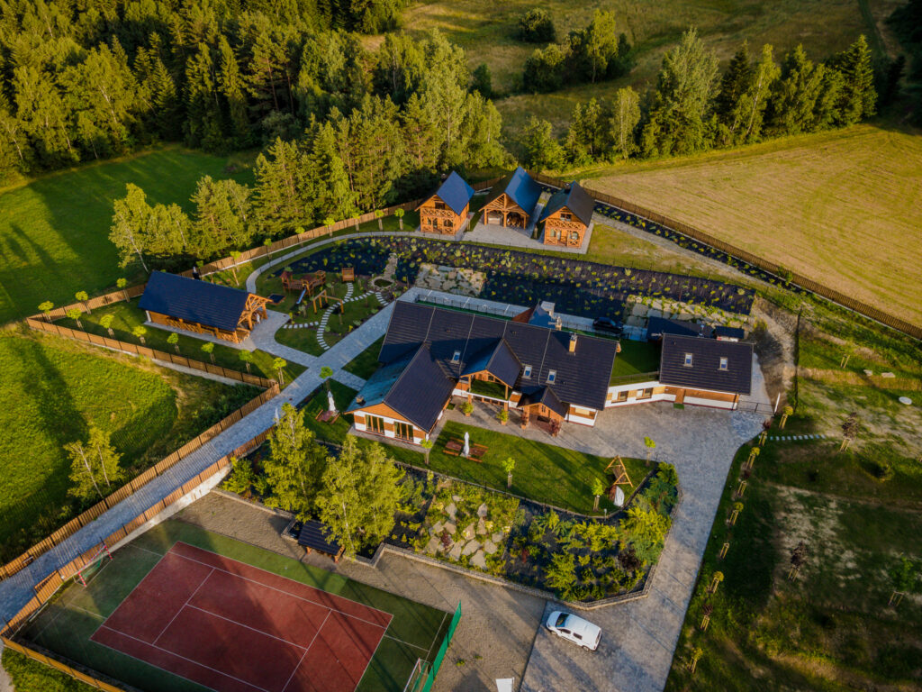 Zdjęcie lotnicze przedstawiające nowoczesny ośrodek Serce Beskidu w Słopnicach, który stanowi idealną bazę wypadową, jeśli interesują Cię ciekawe miejsca w Beskidzie Wyspowym. Na pierwszym planie widać profesjonalny kort tenisowy, a dalej główny budynek z ciemnym dachem, zadbany ogród z alejkami, plac zabaw dla dzieci oraz dwa drewniane domki noclegowe otoczone gęstym lasem i zielonymi polanami.
