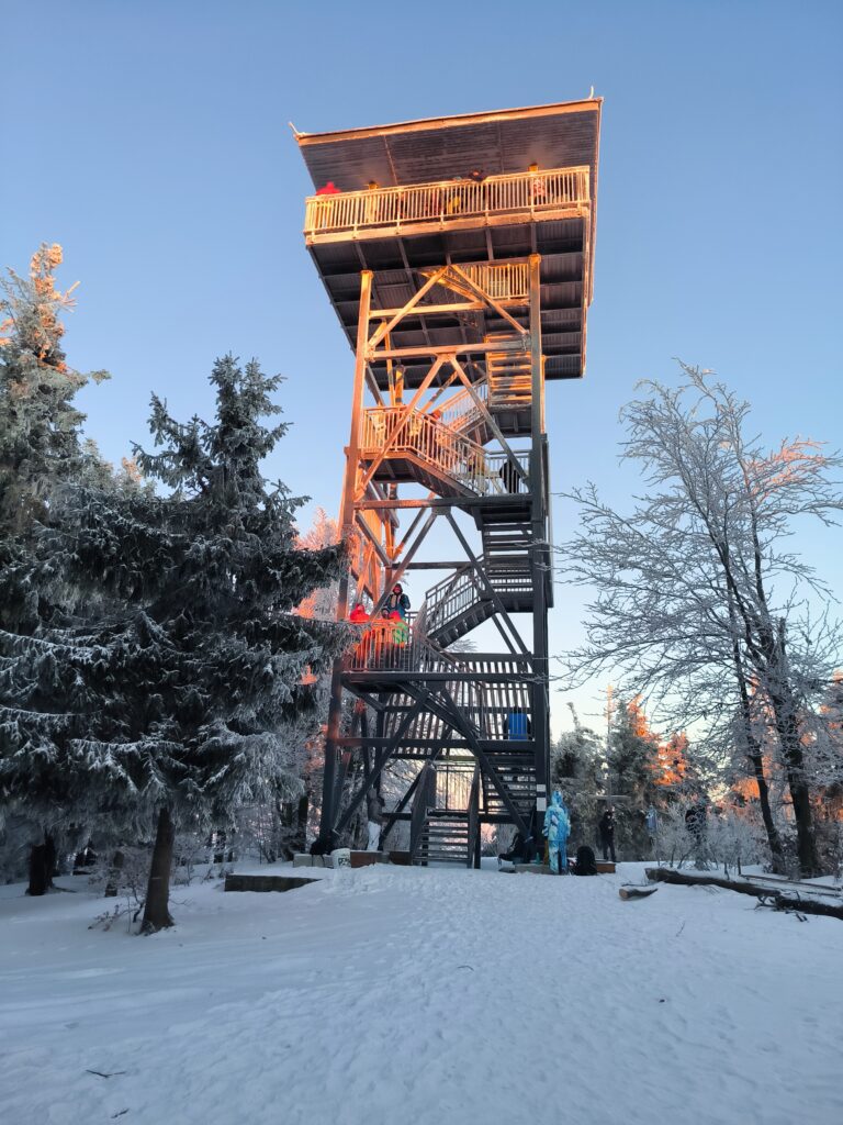 Wysoka, drewniano-metalowa wieża widokowa na szczycie Mogielicy w zimowej szacie, oświetlona promieniami wschodzącego słońca. Wokół konstrukcji rosną drzewa iglaste pokryte grubą warstwą szronu i śniegu, a na platformach widać turystów podziwiających panoramę. Obiekt Serce Beskidu leży u podnóża tej góry, co sprawia, że ta największa atrakcja Beskidu Wyspowego jest dostępna na wyciągnięcie ręki.