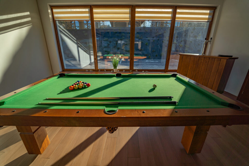 Profesjonalny stół bilardowy z zielonym suknem, ustawiony w jasnym pomieszczeniu z dużym przeszkleniem wychodzącym na teren rekreacyjny Serca Beskidu. Dostęp do gier barowych to sprawdzony sposób na wieczorną rozrywkę, dzięki której integracja zespołu odbywa się poprzez zdrową rywalizację i wspólną zabawę.
