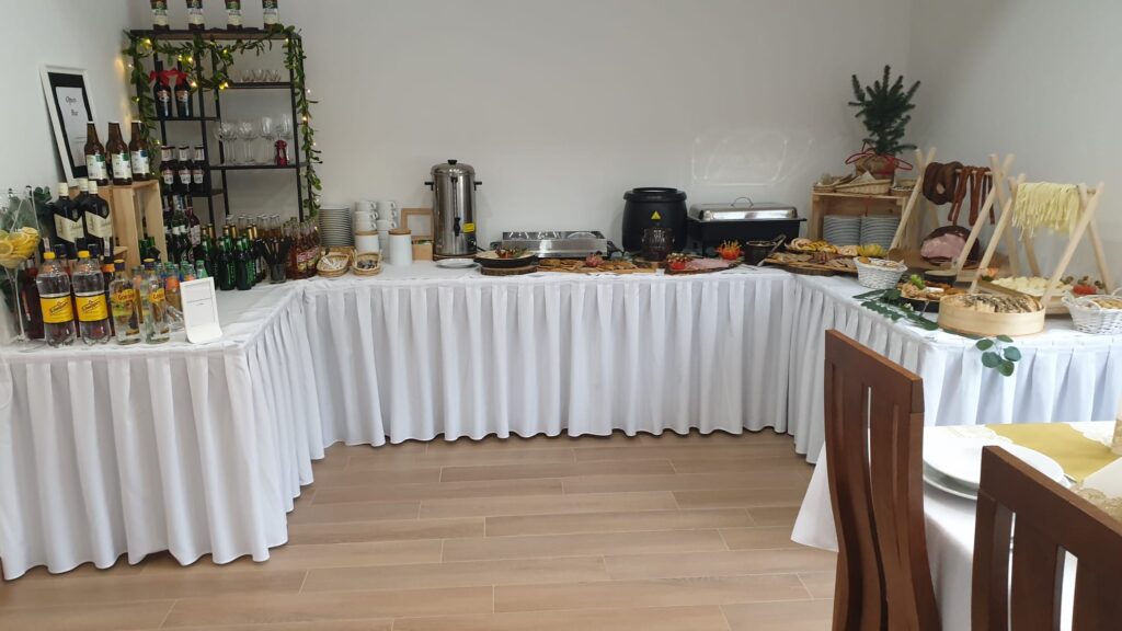 Bufet cateringowy z białym obrusem, bogato zastawiony zimnymi i ciepłymi przekąskami, idealny na spotkanie dla samorządu w centrum konferencyjno-hotelowym Serce Beskidu w Słopnicach w Małopolsce. Widoczne lokalne sery, wędliny, owoce, napoje oraz bufet kawowy.