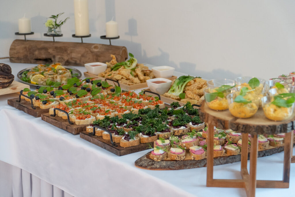Zbicie ujęcie elegancko zaaranżowanego bufetu z cateringiem na spotkanie biznesowe. Widoczne są różnorodne, estetycznie podane przekąski typu finger food, w tym bruschetty z pomidorami i bazylią, kanapeczki z ciemnego pieczywa z pastami i zieleniną oraz smażone kąski. Dania są prezentowane na drewnianych deskach i pieńkach, a w tle widać minimalistyczne dekoracje ze świec. Tego typu wysokiej jakości catering jest dostępny przy wynajem sali konferencyjnej w górach w hotelu Serce Beskidu.