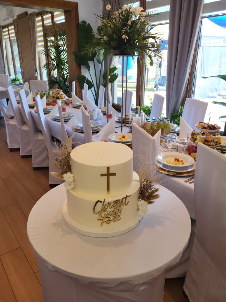 Elegancki, biały tort piętrowy z napisem "Chrzest Święty Poli" oraz złotym krzyżem, przygotowany na przyjęcie w Sercu Beskidu. W tle widać długi stół z zastawą i dekoracjami roślinnymi. Chrzciny to wyjątkowo intymna impreza okolicznościowa, którą w naszym obiekcie celebrujemy z dbałością o najmniejszy szczegół.