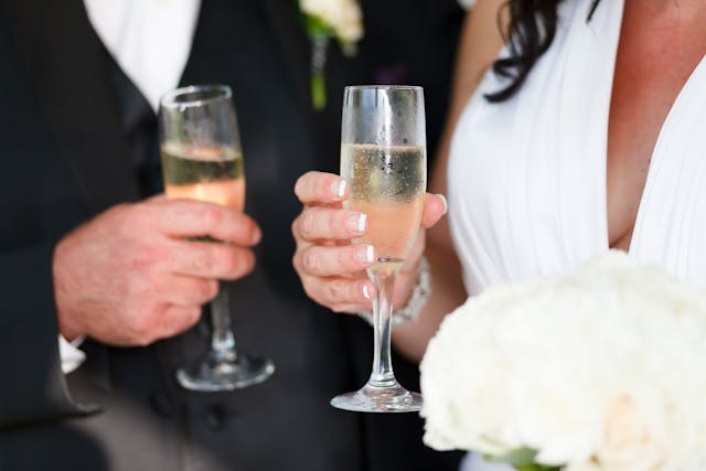 Zbliżenie na dłonie Pary Młodej (lub gości) wznoszących toast dwoma kieliszkami szampana. Kobieta ma elegancki biały manikiur, a mężczyzna ma na sobie czarny garnitur, co jest przykładem, Jak się ubrać na góralskie wesele jako gość.