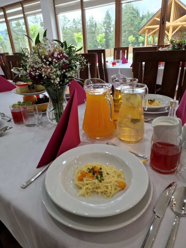 Zbliżenie na nakrycie stołu podczas bankietu firmowego w górach w centrum konferencyjnym Serce Beskidu w Słopniach. Na białym obrusie widoczny jest talerz z makaronem, różowe serwetki, karafki z sokami oraz świeże kwiaty w wazonie, co podkreśla elegancki bankiet firmowy i dbałość o detale.