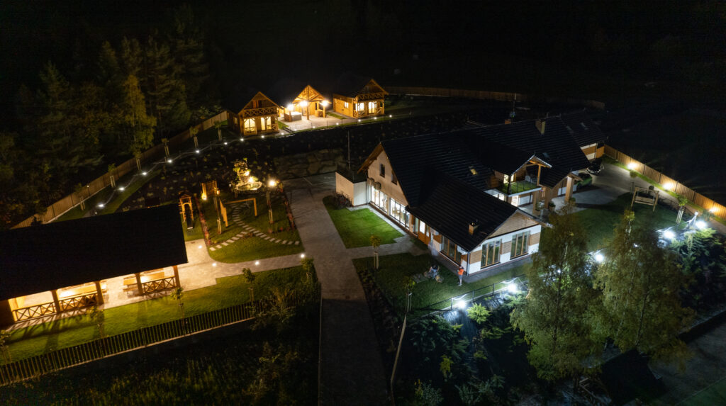 Widok z lotu ptaka na oświetlony nocą obiekt Serce Beskidu. Na zdjęciu widać duży budynek główny z przeszkloną salą, dwa drewniane domki regionalne oraz altanę grillową, co pokazuje jak wygląda profesjonalna organizacja imprezy firmowej w górskim otoczeniu.
