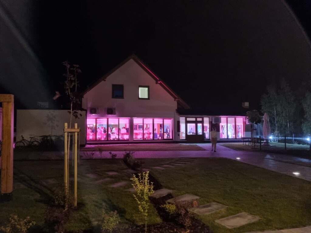 Zewnętrzny widok na budynek Serce Beskidu po zmroku. Przeszklona sala bankietowa na parterze jest intensywnie podświetlona na kolor fuksji/różowy, a przez okna widać bawiących się wewnątrz gości. Przed budynkiem znajduje się zadbany trawnik z młodymi nasadzeniami i ścieżką z płyt kamiennych. Nowoczesna bryła obiektu z jasną elewacją i dwuspadowym dachem prezentuje się niezwykle atrakcyjnie jako miejsce na profesjonalnie zorganizowane wydarzenie wyjazdowe.