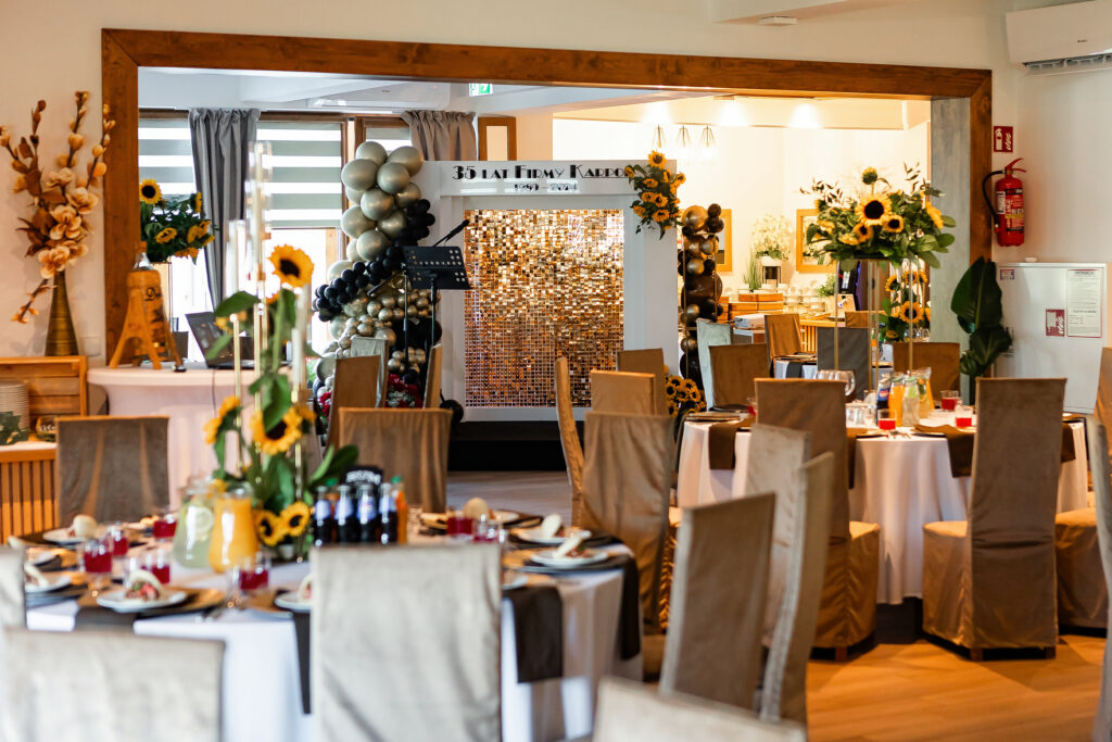 Sala bankietowa w Sercu Beskidu przygotowana na jubileusz 35-lecia firmy. Widoczne są okrągłe stoły z dekoracjami ze słoneczników oraz złota ścianka dekoracyjna z balonami, co pokazuje, jak zorganizować bankiet o prestiżowym charakterze.