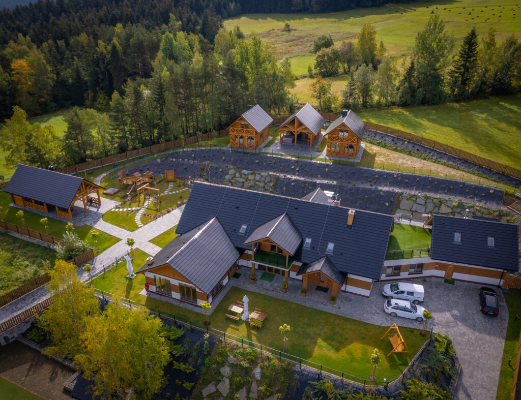 Szerokie, panoramiczne ujęcie z drona obrazujące cały, luksusowy kompleks rekreacyjno-weselny Serce Beskidu w Słopnicach, w otoczeniu malowniczych wzniesień Beskidu Wyspowego. Na pierwszym planie dominuje duży, reprezentacyjny budynek główny z białym tynkiem, drewnianym wykończeniem elewacji oraz wielospadowym, ciemnym dachem z oknami połaciowymi. Przed budynkiem rozpościera się perfekcyjnie utrzymany, zielony trawnik, na którym ustawiono drewniane meble biesiadne z białym parasolem oraz drewnianą huśtawkę ogrodową dla najmłodszych. Po prawej stronie widać wybrukowany parking z zaparkowanymi samochodami osobowymi. Powyżej, na kaskadowo ułożonym terenie, wznoszą się trzy drewniane domki noclegowe oraz otwarta, zadaszona altana grillowa. W tle rozciągają się rozległe, zielone polany z tradycyjnymi kopkami siana, pasma drzew liściastych i iglastych oraz łagodne szczyty górskie, co wizualnie potwierdza, dlaczego warto wybrać to miejsce na wyjątkowe wesele w górach z noclegiem.