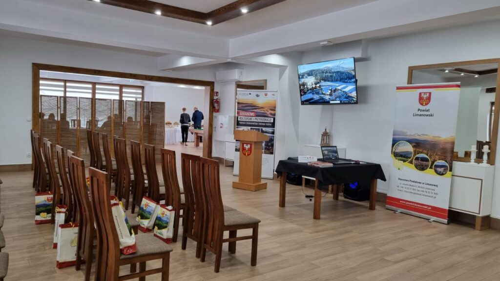 Widok na salę konferencyjną w układzie kinowym, przygotowaną na dużą prezentację lub wydarzenie dla instytucji w górach w obiekcie Serce Beskidu. Widoczne rzędy drewnianych krzeseł, podium, roll-upy informacyjne oraz duży telewizor ścienny.