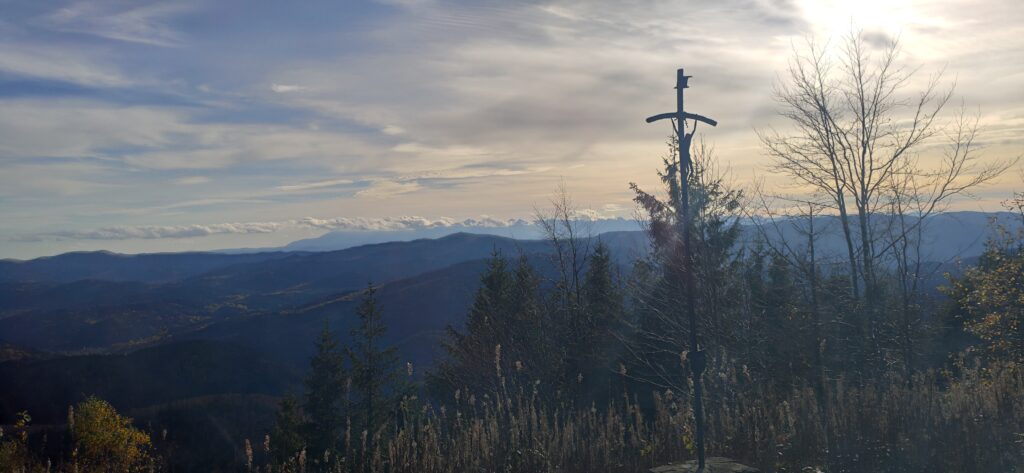 Wysoki, metalowy krzyż o ażurowej konstrukcji, stojący na kamiennym postumencie na szczycie góry. Tło stanowi złota godzina zachodu słońca nad pasmami górskimi. Ważny przystanek dla zdobywców odznaki Korona Beskidu Wyspowego, na Mogielicy, u podnóża której znajduje się ośrodek Serce Beskidu.