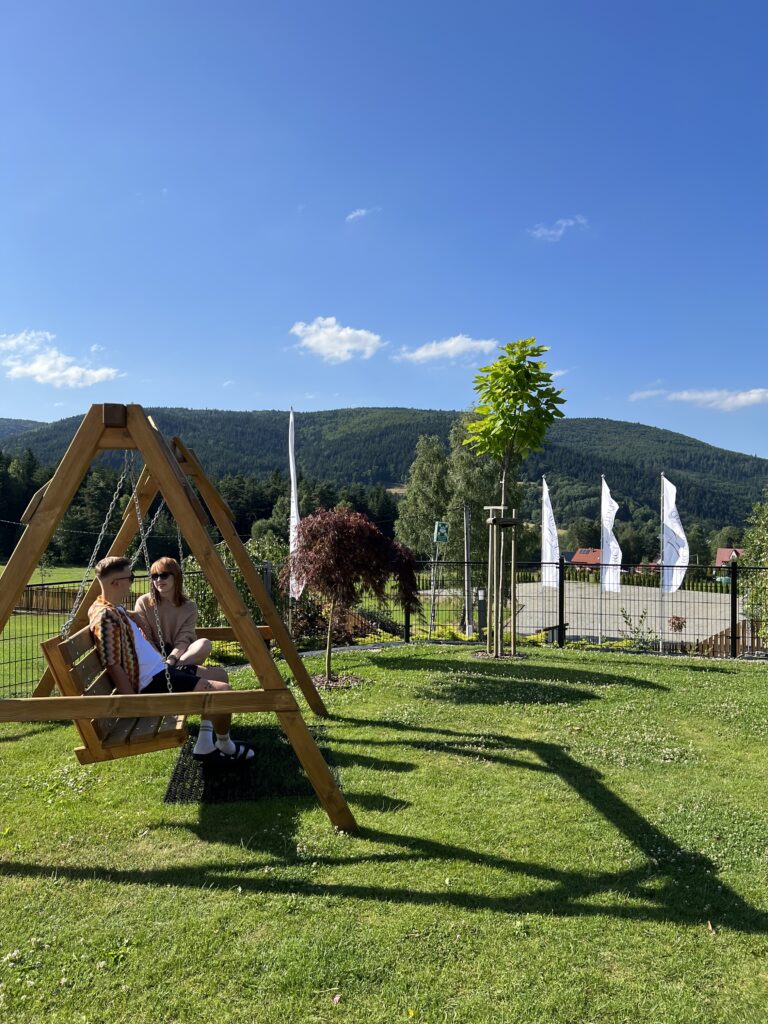 Para pracowników rozmawia na dużej drewnianej huśtawce na zadbanym trawniku w Sercu Beskidu, mając przed sobą panoramę zielonych szczytów. Taki wyjazd integracyjny w otoczeniu natury pozwala pracownikom wyjść z ról służbowych i poznać się lepiej.