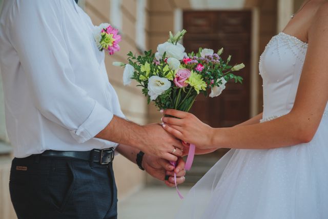 Zbliżenie na Pannę Młodą i Pana Młodego stojących naprzeciw siebie i wspólnie trzymających bukiet ślubny przed wejściem do obiektu Serce Beskidu. Kobieta po prawej stronie ubrana jest w białą suknię ślubną z gorsetem zdobionym drobnymi perełkami i tiulowym dołem. Mężczyzna po lewej stronie ma na sobie elegancką, białą koszulę z podwiniętymi rękawami, ciemne spodnie z czarnym paskiem oraz dopasowaną do bukietu butonierkę z różowo-zielonych kwiatów wpiętą w pierś. Ich dłonie splatają się na łodygach bukietu ślubnego, w którym dominują białe eustomy, jasnoróżowe róże oraz zielone gałązki, przewiązane subtelną, satyną tasiemką w kolorze różowym. Na palcu mężczyzny widoczna jest złota obrączka ślubna. W rozmytym tle widać fragment beżowej elewacji oraz duże, ciemnobrązowe drzwi wejściowe. Fotografia idealnie oddaje romantyczny klimat uroczystości, jaką jest kompleksowo zorganizowane wesele w górach z noclegiem.