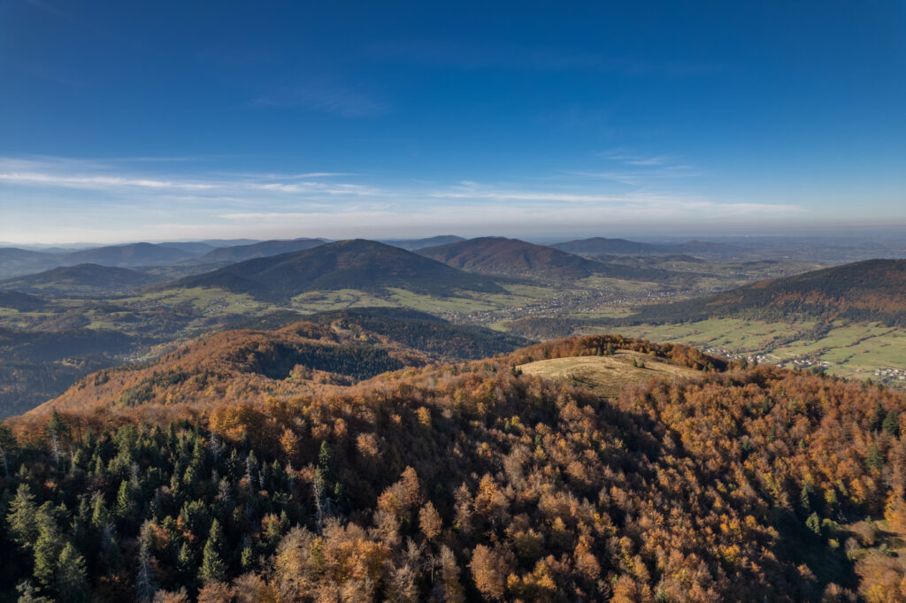 Lotnicze ujęcie przedstawiające górzysty krajobraz z wyraźną dominacją zielonych lasów iglastych i liściastych. W oddali widać charakterystyczne, odosobnione wzniesienia wyłaniające się z mgieł, które definiują Beskid Wyspowy. W jednej z widocznych dolin u podnóża gór znajduje się ośrodek Serce Beskidu.