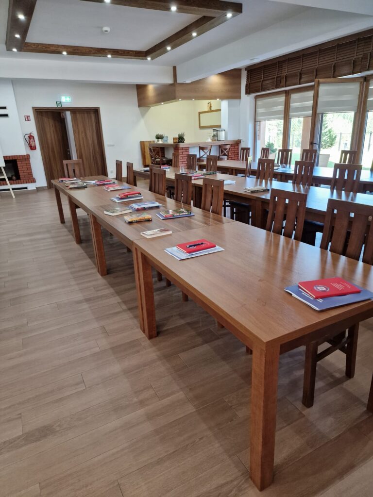 Nowoczesna, jasna sala szkoleniowa z długimi, drewnianymi stołami i krzesłami, przygotowana do przyjęcia uczestników. Na stołach leżą materiały w czerwonych okładkach, idealne do organizacji wydarzenie dla pracowników instytucji lub dużej konferencji w centrum szkoleniowo-konferencyjnym Serce Beskidu w górach Beskidach.