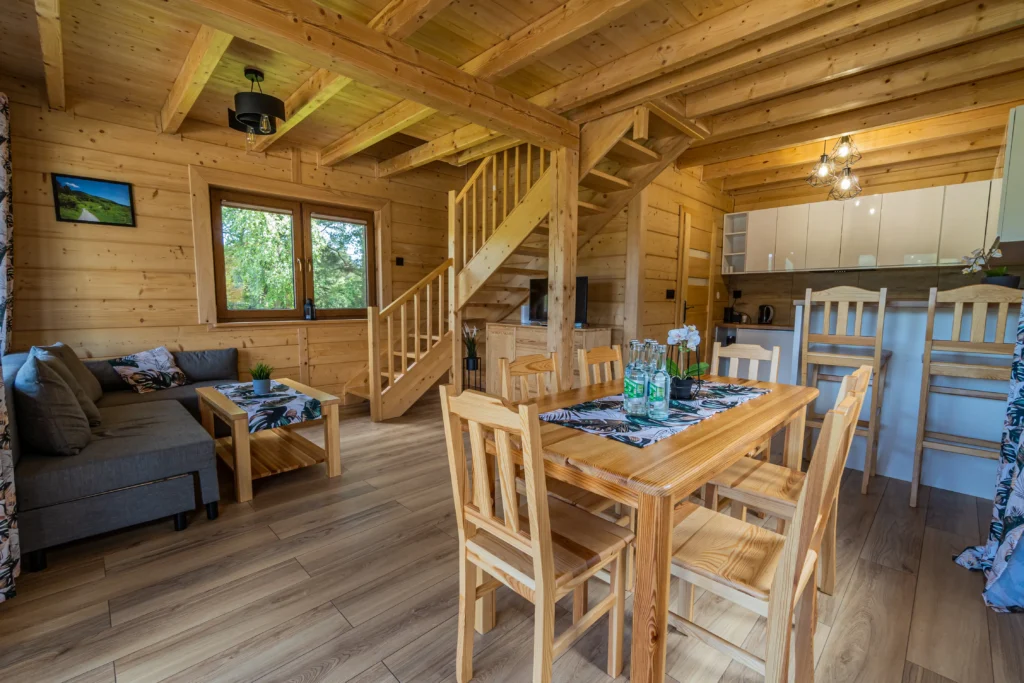 Wnętrze luksusowego domku z bali w ośrodku Serce Beskidu. Widoczny drewniany salon z szarą kanapą, stołem jadalnianym, schodami na piętro oraz w pełni wyposażonym aneksem kuchennym, idealny jako baza noclegowa na warsztaty rozwoju osobistego.