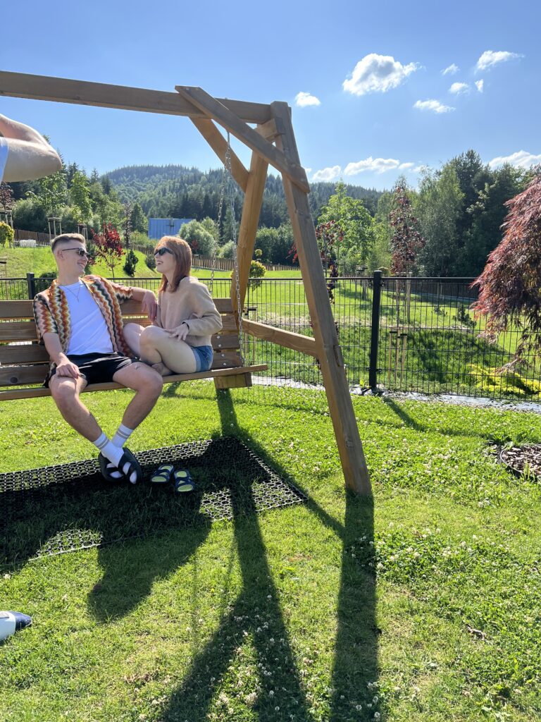 Dwoje uśmiechniętych uczestników wyjazdu firmowego odpoczywa na dużej, drewnianej huśtawce w ogrodzie obiektu Serce Beskidu, ciesząc się słoneczną pogodą i widokiem na góry. Taka swobodna atmosfera sprzyja budowaniu więzi, sprawiając, że impreza firmowa w górach staje się autentycznym doświadczeniem łączącym zespół.