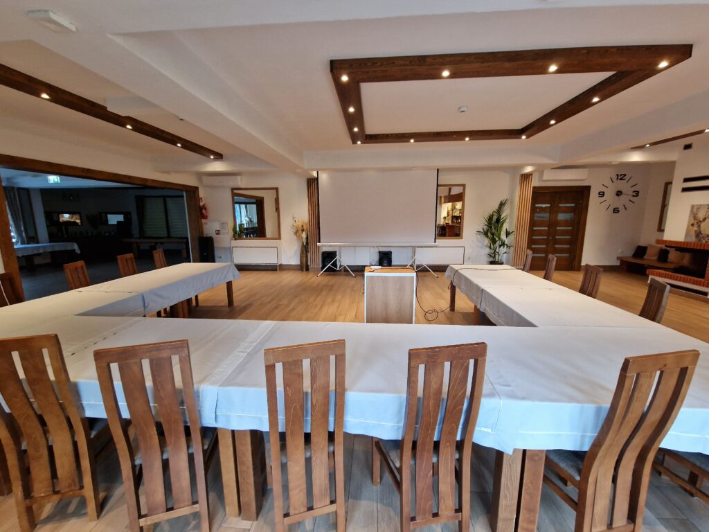 Nowoczesna i jasna sala konferencyjna w górach w obiekcie Serce Beskidu urządzona w układzie „U”, z długimi stołami przykrytymi jasnym obrusem i drewnianymi, wyprofilowanymi krzesłami. W centralnej części sali znajduje się mównica i duży ekran projekcyjny, a drewniane elementy konstrukcyjne sufitu dodają wnętrzu górskiego, eleganckiego charakteru. Jest to elastyczna przestrzeń do organizacji spotkań biznesowych i szkoleń.