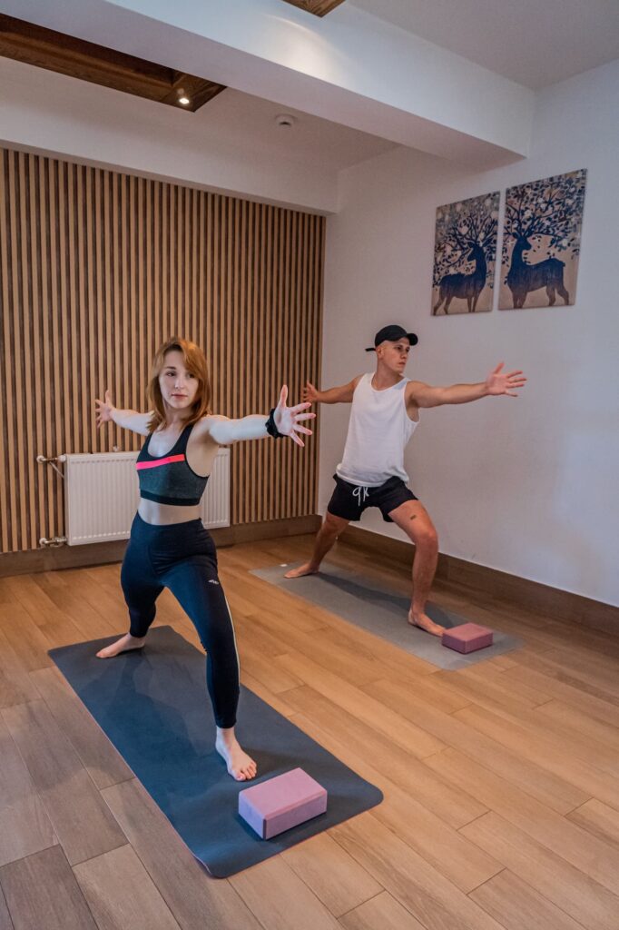 Kobieta i mężczyzna w pozycji Wojownika II podczas sesji w sali warsztatowej. Wnętrze posiada nowoczesne lamele na ścianach, drewnianą podłogę i klimatyczne obrazy z jeleniami, tworząc profesjonalne warunki na warsztaty z jogą i rozwojem osobistym w górach Beskidu Wyspowego w Słopnicach.