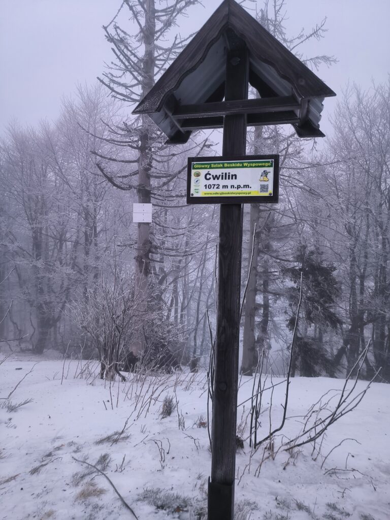 Zbliżenie na drewniany drogowskaz z tabliczką „Ćwilin 1072 m n.p.m.” oznaczającą szlak Beskidu Wyspowego w zimowej, mglistej scenerii. Widoczne oszronione drzewa i śnieg pokrywający polanę tworzą surowy, górski klimat, który można poczuć podczas wypraw organizowanych z bazy Serce Beskidu.