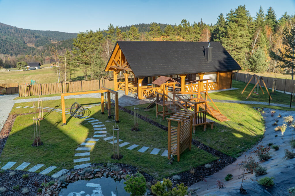 Widok z góry na duży, drewniany plac zabaw dla dzieci oraz altanę grillową w Serce Beskidu, otoczony zielenią i panoramą Beskidu Wyspowego, co pomaga ocenić, co jest lepsze góry czy plaża dla rodzinnej rekreacji z dziećmi.