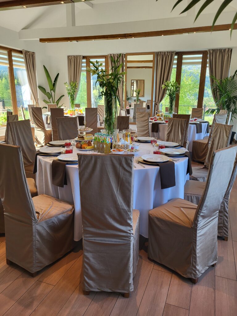 Widok na okrągły stół bankietowy w jasnej sali Serca Beskidu z wysokimi krzesłami w pokrowcach. Na stole znajdują się napoje, owoce i wysokie wazony z kwiatami, co stanowi doskonały przykład na elegancki bankiet dla kontrahentów.