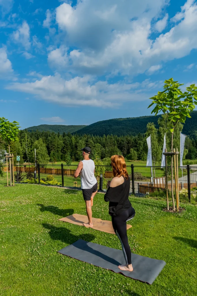 Grupa osób praktykuje jogę na zielonym trawniku przed ośrodkiem Serce Beskidu w Słopnicach. Uczestnicy wykonują asany balansujące na matach, otoczeni naturą i widokiem na zalesione szczyty Beskidu Wyspowego pod błękitnym niebem.