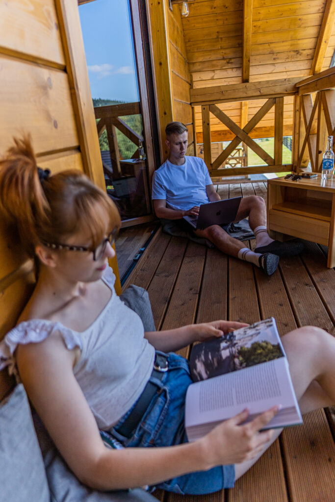 Na tarasie obiektu Serce Beskidu kobieta w okularach czyta album, siedząc na drewnianej podłodze, podczas gdy w tle mężczyzna pracuje na laptopie. To zdjęcie idealnie obrazuje, że wyjazd integracyjny to przestrzeń na swobodne budowanie relacji i odpoczynek od biurowego pędu.