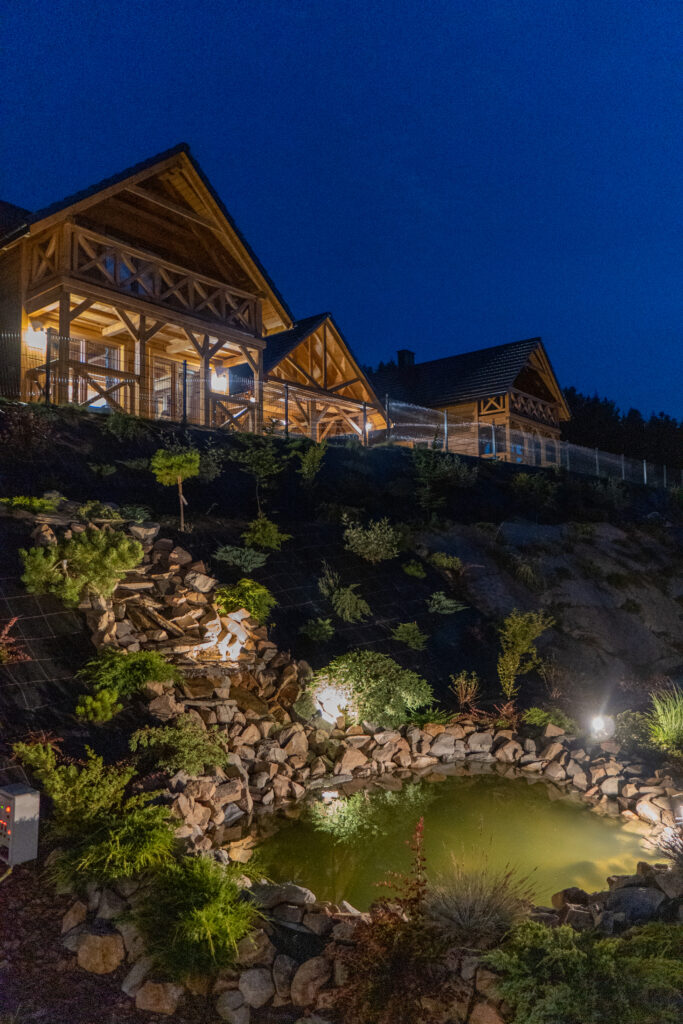 Nocne ujęcie oświetlonych, drewnianych domków w Sercu Beskidu, położonych na wzgórzu nad ozdobnym stawem z kaskadą kamienną, co tworzy klimat nieosiągalny podczas dylematu wyjazd w góry czy nad morze.