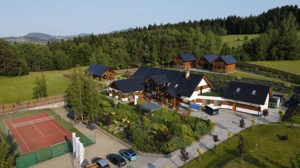 Widok na budynek główny oraz dwa sąsiadujące, drewniane domki całoroczne w Sercu Beskidu. W dolnej części zdjęcia widoczny fragment kortu tenisowego oraz zaparkowane samochody. To miejsce łączy funkcję pracy i wypoczynku jako ośrodek konferencyjny w górach.
