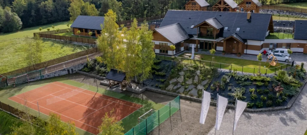 Zdjęcie z lotu ptaka przedstawiające ośrodek Serce Beskidu, składający się z sali weselnej/hotelu i domków noclegowych w górskim, drewnianym stylu. Otoczenie jest zielone, z widocznym kortem tenisowym i zadbanym ogrodem, co pokazuje, Gdzie zorganizować modne wesele w 2026 zapewniające atrakcje plenerowe i noclegi.
