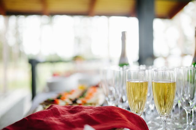 Bufet z rzędem kieliszków wypełnionych musującym napojem (prosecco/szampan) i butelką w tle. W oddali widać bufet z jedzeniem. Wybór alkoholu jest kluczowy dla ceny za talerzyk wesele 2026.
