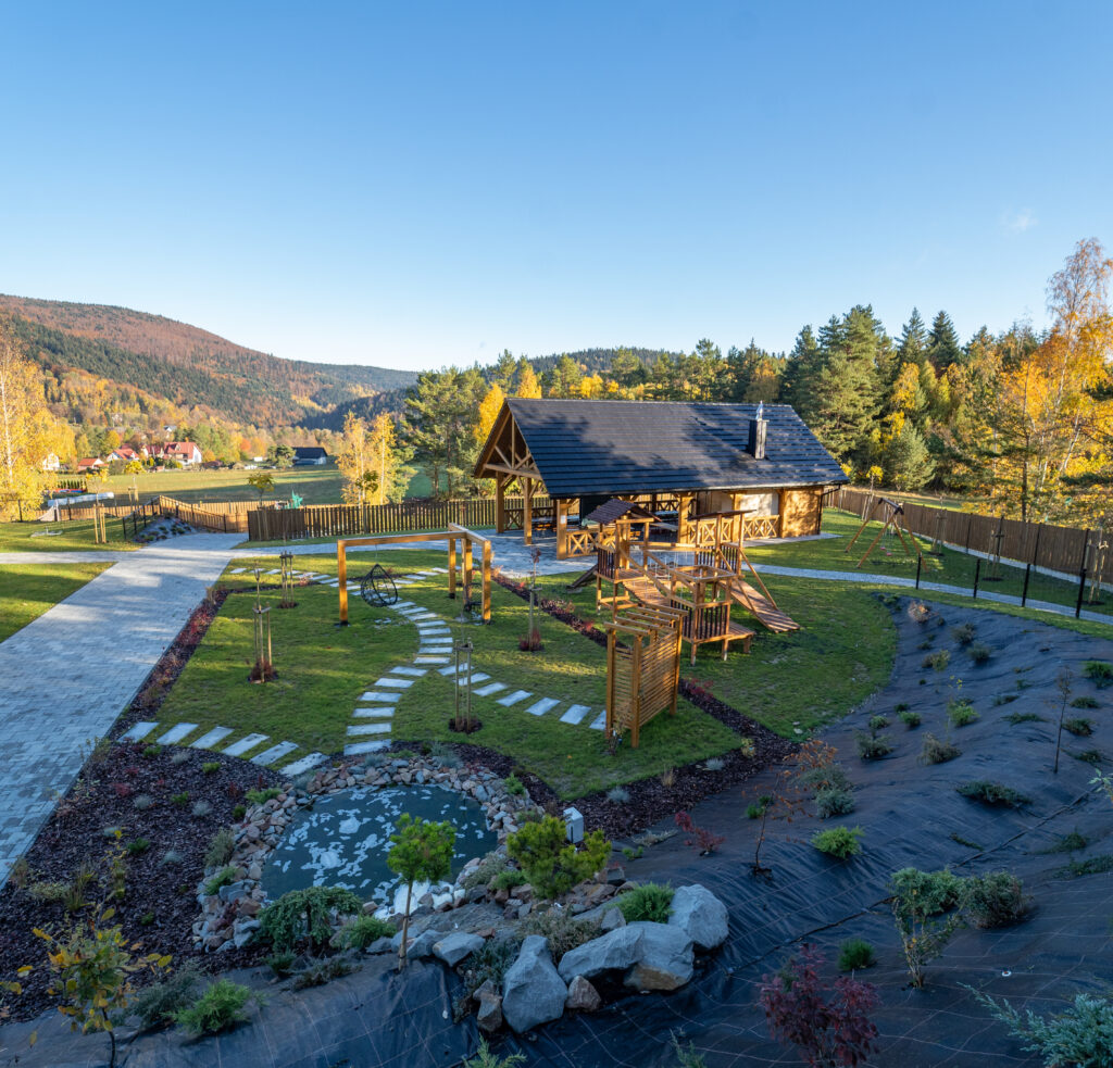 Zdjęcie przedstawiające zagospodarowany teren rekreacyjny ośrodka w górach, z widokiem na altanę grillową (zadaszony budynek z drewna) i Przed altaną znajduje się trawiasty teren z kamienną ścieżką w kształcie liścia i małe oczko wodne otoczone kamieniami. Otoczenie w pełnej krasie jesieni, z drzewami w odcieniach złota i czerwieni. Infrastruktura ta świetnie uzupełnia ofertę dla firm organizujących firmowy wyjazd w Beskidy do hotelu Serce Beskidu w Słopnicach.