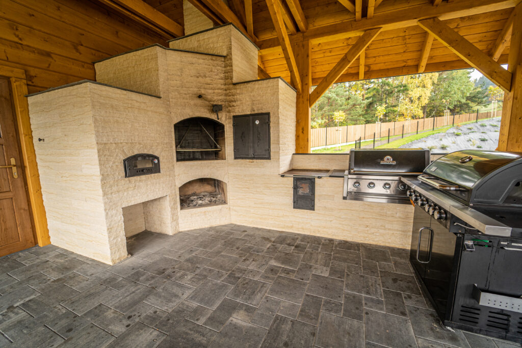 Szczegółowe ujęcie przestronnej, zadaszonej altany grillowej w górskim hotelu, wykonanej z naturalnego, jasnego drewna, z kamiennym kominkiem, tradycyjnym piecem opalanym drewnem i nowoczesnym, czarnym grillem gazowym; idealne miejsce na firmowy wieczór wigilijny lub ognisko integracyjne w hotelu Serce Beskidu w Słopnicach.