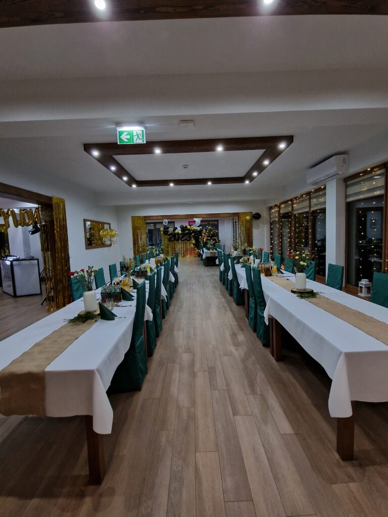 Przestronna sala w obiekcie Serce Beskidu przygotowana na przyjęcie. Długie stoły nakryte białymi obrusami z jutowymi bieżnikami i zielonymi akcentami tworzą elegancki klimat. W tle widoczne są świąteczno-noworoczne dekoracje balonowe, co ilustruje profesjonalną organizację imprez firmowych świątecznych w górach Beskidach w Serce Beskidu.