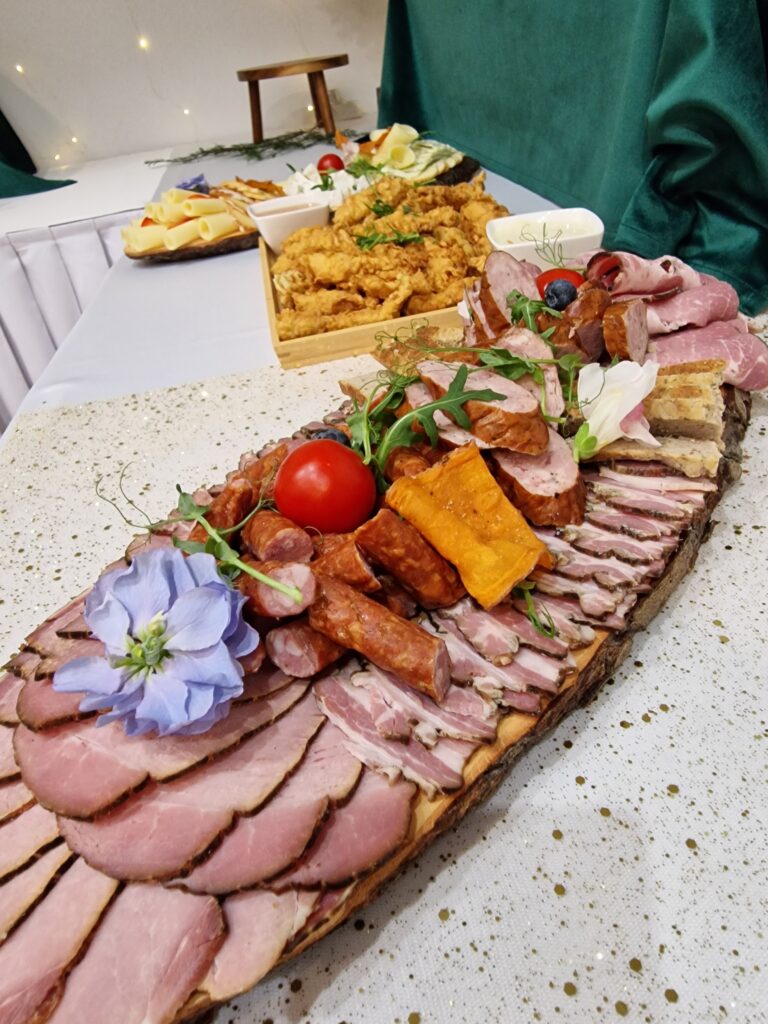 Zbliżenie na drewnianą deskę wypełnioną swojskimi wędlinami, kiełbasami i pieczonym mięsem, udekorowaną świeżymi ziołami i kwiatami. W tle widać kolejne półmiski z przekąskami, co jest doskonałym przykładem menu na tradycyjne wigilie firmowe czerpiące z lokalnych smaków Beskidu.