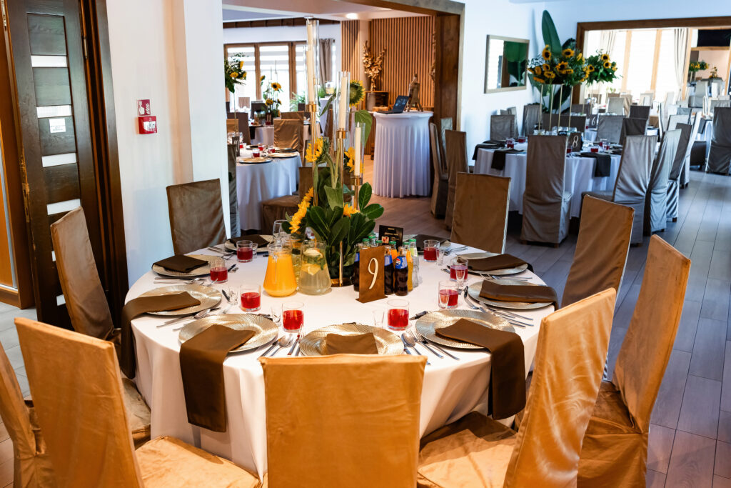 Przygotowana, elegancka sala restauracyjna w hotelu Serce Beskidu w Beskidzie Wyspowym z okrągłymi stołami na bankiet, z brązowo-złotymi pokrowcami na krzesła. Stół na pierwszym planie jest nakryty białym obrusem, brązowymi serwetami i talerzami z podkładkami. Centralnym elementem dekoracji są słoneczniki, wysokie świece i dzbanek z lemoniadą. W tle widoczne kolejne stoły przygotowane na spotkanie firmowe. Restauracja Serce Beskidu w Słopnicach to idealne miejsce na firmowy event, na który można założyć modną stylizację na firmową wigilię w górach Beskidach.