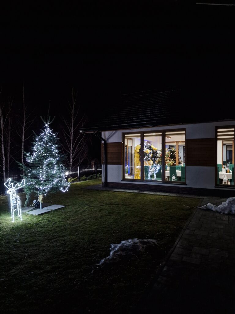 Nocne ujęcie budynku Serce Beskidu z zewnątrz. Na trawniku stoi podświetlana ledowa figura renifera oraz choinka z lampkami. Przez duże okna widać oświetlone wnętrze sali bankietowej z gośćmi, co pokazuje, jak efektownie mogą wyglądać trendy w organizacji firmowych imprez świątecznych 2025.