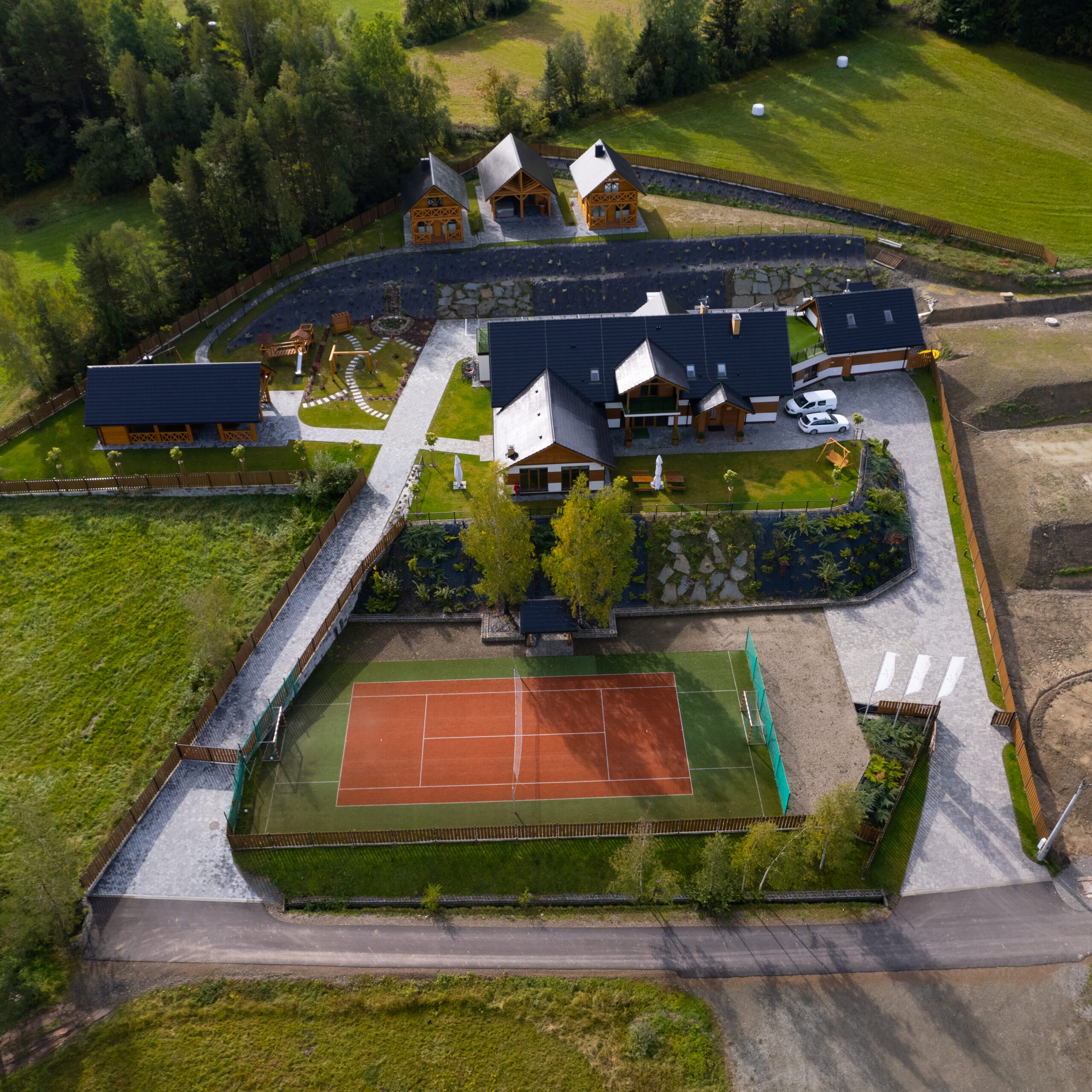 Zdjęcie z drona ukazujące pełną infrastrukturę kompleksu Serce Beskidu, w tym kort tenisowy, budynek główny, domki oraz tereny zielone otoczone lasem. Rozległy teren i bogate zaplecze sportowo-rekreacyjne czynią ten obiekt doskonałą lokalizacją na udane imprezy integracyjne dla wymagających firm.