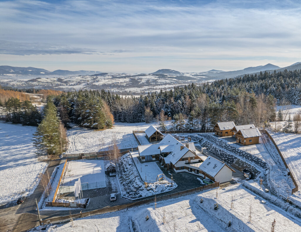 Panorama zimowego krajobrazu Beskidu Wyspowego z lotu ptaka, przedstawiająca kompleks hotelowy Serce Beskidu z budynkami o jasnych elewacjach i białych dachach pokrytych śniegiem, otoczony zaśnieżonym lasem i górami, idealna atrakcja wigilię firmową połączoną z wyjazdem integracyjnym w góry Beskidy.