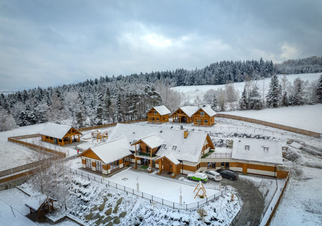 Ujęcie z drona przedstawiające kompleks wypoczynkowy hotelu Serce Beskidu w Słopnicach zimą. Wszystkie dachy pokryte są śniegiem, otoczenie stanowią zaśnieżone pola i gęsty, ośnieżony las. Widoczne są budynki główne oraz mniejsze domki rekreacyjne, ogrodzony teren oraz podjazd, co świadczy o bogatej infrastrukturze na wigilia firmowa w górach Beskidach w hotelu Serce Beskidu.