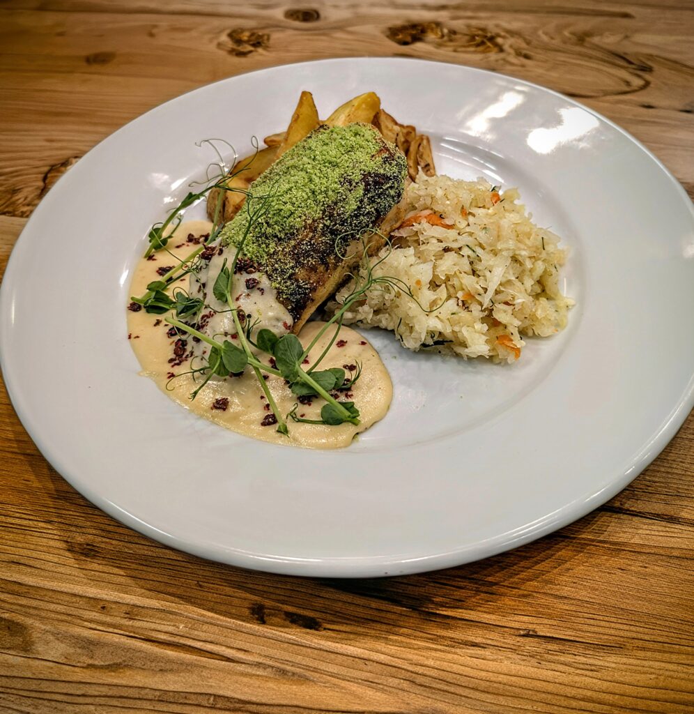 Wykwintnie podane danie główne na okrągłym, białym talerzu, idealne jako rybna propozycja w menu na wigilię firmową w restauracji Serce Beskidu w Słopnicach w górach Beskidu Wyspowego. Składa się z kawałka ryby (prawdopodobnie dorsza lub sandacza) pokrytego zieloną posypką (prawdopodobnie zioła), podanego na kremowym, jasnym sosie z dodatkiem czerwonych przypraw i kiełków. Towarzyszy mu porcja kiszonej kapusty oraz pieczone ziemniaki.