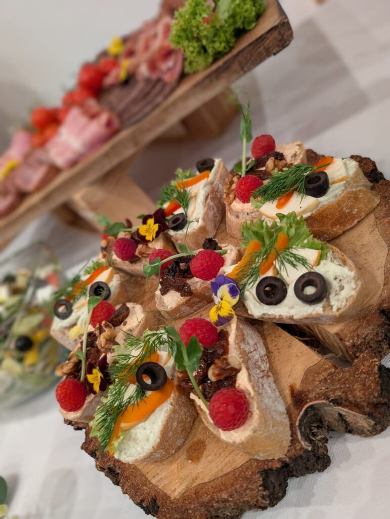 Zbliżenie na okrągłą, drewnianą tacę z korą, na której ułożono mini kanapki koktajlowe z różnymi pastami, idealne jako część cateringu na wigilia firmowa menu. Dekorowane są one malinami, orzechami włoskimi, marchewką, oliwkami oraz jadalnymi kwiatami, tworząc elegancki bufet zimnych przekąsek serwowanych na spotkaniu świątecznym w hotelu Serce Beskidu w Małopolsce, w górach Beskidach.
