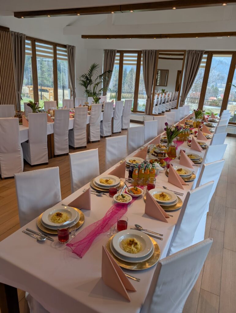 Szerokie ujęcie eleganckiej, jasnej sali restauracyjnej z dużymi panoramicznymi oknami, z widokiem na góry, przystosowanej do zorganizowania wigilia firmowa w restauracji. Długi stół bankietowy jest nakryty białym obrusem, różowym bieżnikiem oraz eleganckimi zastawami z podtalerzami w kolorze złotym. Krzesła są ubrane w białe pokrowce.