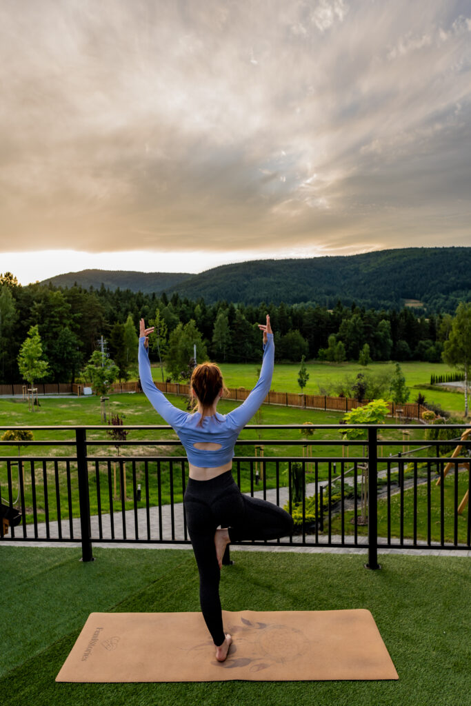 Kobieta o rudych włosach stoi tyłem do obiektywu w pozycji Drzewa (Vrksasana) na macie na tarasie widokowym Serce Beskidu o zachodzie słońca. W tle malowniczy widok na Beskidy. Taki wyjazd integracyjny w góry to świetny prezent firmowy na święta dla budowania relacji.