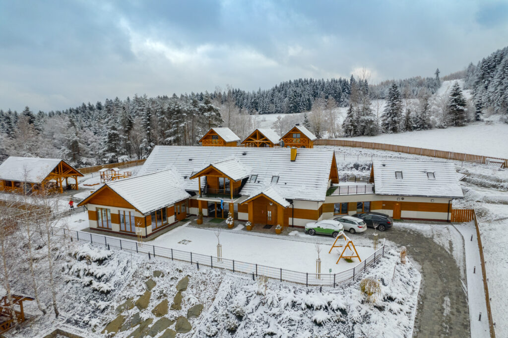 Widok z lotu ptaka na ośrodek konferencyjny Serce Beskidu w Słopnicach przykryty grubą warstwą śniegu, co tworzy magiczny klimat na wyjazd integracyjny w Beskidy. Fotografia prezentuje główny budynek z charakterystycznymi drewnianymi elementami elewacji, trzy mniejsze domki góralskie w tle oraz plac zabaw i parking z samochodami. Całość otoczona jest gęstym, zaśnieżonym lasem iglastym, co podkreśla unikalną lokalizację obiektu na event firmowy w górach Małopolski organizowany z okazji podsumowania roku w firmie.