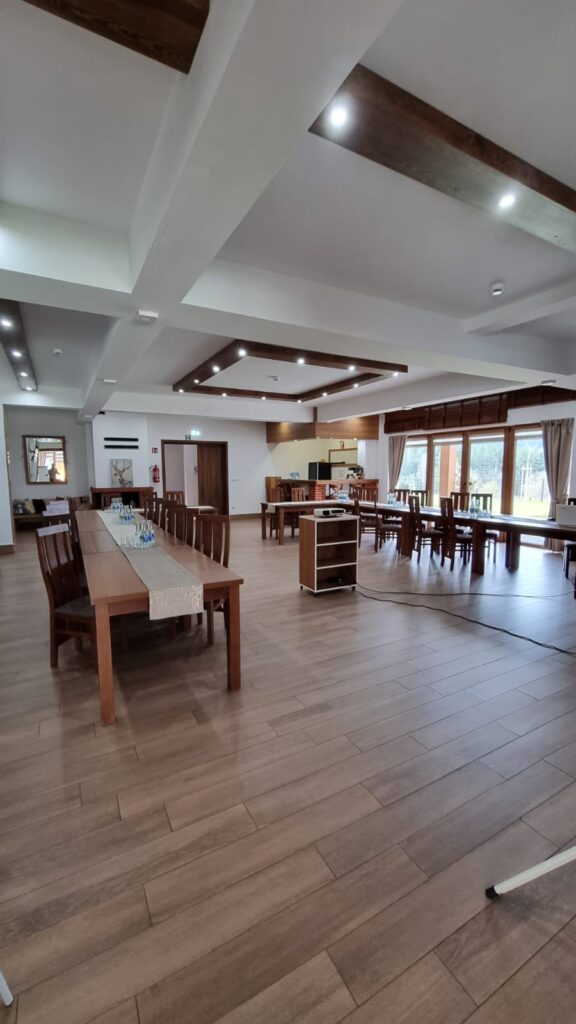 Wnętrze przestronnej sali w ośrodku Serce Beskidu z drewnianą podłogą i sufitem zdobionym belkami, gdzie zorganizowano podsumowanie roku dla pracowników. Widoczne są długie, dębowe stoły ustawione w literę U, na których przygotowano wodę mineralną dla uczestników. W głębi znajduje się barek kawowy, nowoczesne oświetlenie LED w kasetonach oraz duże przeszklenia prowadzące na taras, co tworzy idealne warunki na prestiżowy event firmowy.