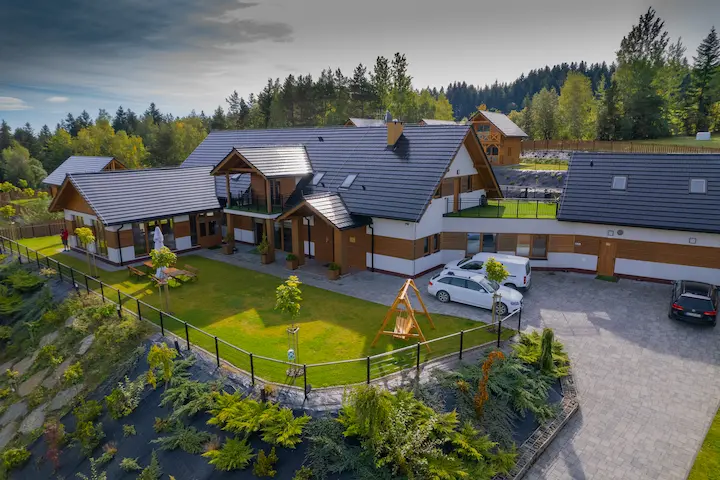 Zdjęcie przedstawiające nowoczesny obiekt eventowy Serce Beskidu w Słopnicach. Widoczna jest architektura budynku z ciemnym dachem, zielony teren rekreacyjny, parking oraz otaczające lasy, co czyni go idealnym miejscem na szkolenie dla grup zorganizowanych z okolic Krakowa.