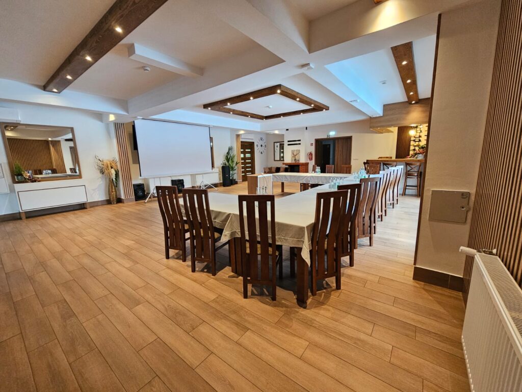 Nowoczesna sala szkoleniowa w Sercu Beskidu przygotowana na biznesowe podsumowanie roku. Na zdjęciu widać duży, biały ekran projekcyjny, profesjonalne kolumny nagłośnieniowe oraz stoły przykryte eleganckimi, jasnymi obrusami. Jasne ściany z elementami lamelowymi oraz stylowy zegar ścienny dopełniają profesjonalny charakter wnętrza, które świetnie sprawdza się jako miejsce na strategiczne spotkanie zarządu z zespołem.