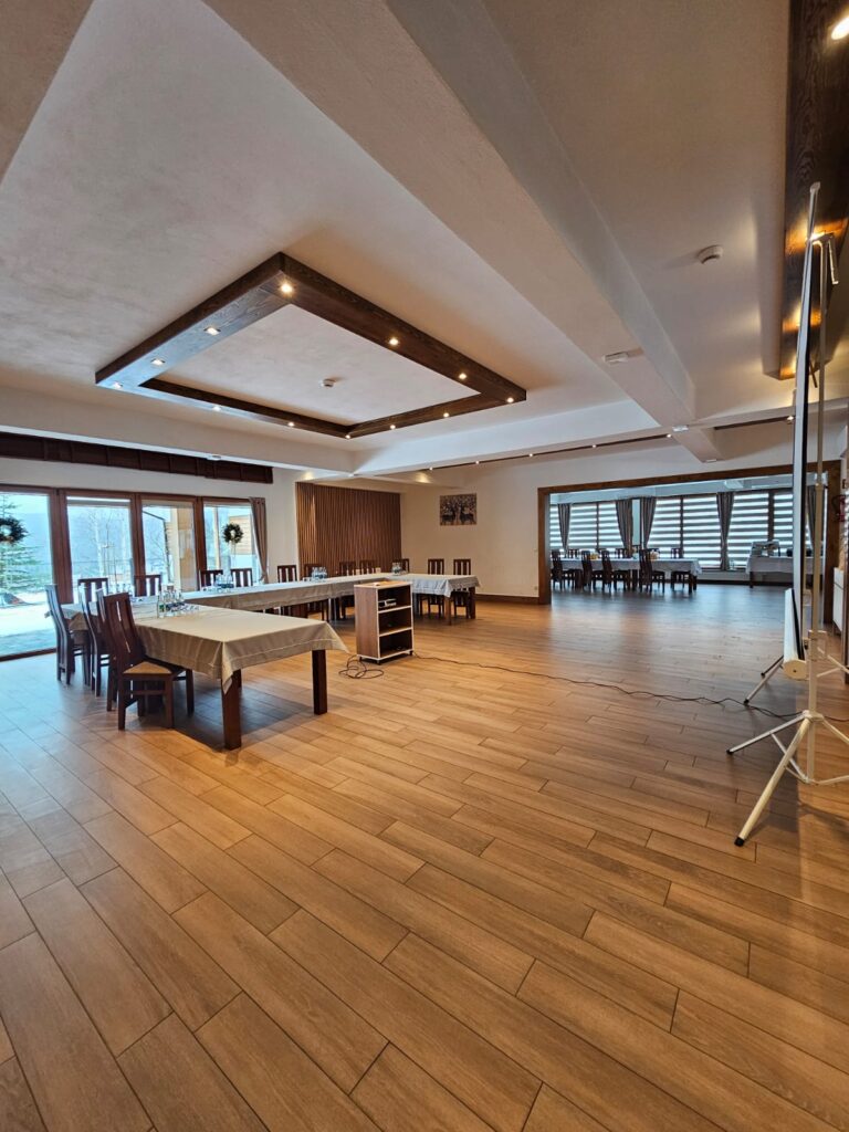 Nowoczesna sala konferencyjna w Beskidach w ośrodku Serce Beskidu, wyposażona w duże okna tarasowe, drewnianą podłogę i profesjonalne oświetlenie sufitowe. Na pierwszym planie widoczne są stoły z jasnymi obrusami ustawione w podkowę, przygotowane na szkolenie dla grup zorganizowanych z okolic Krakowa.