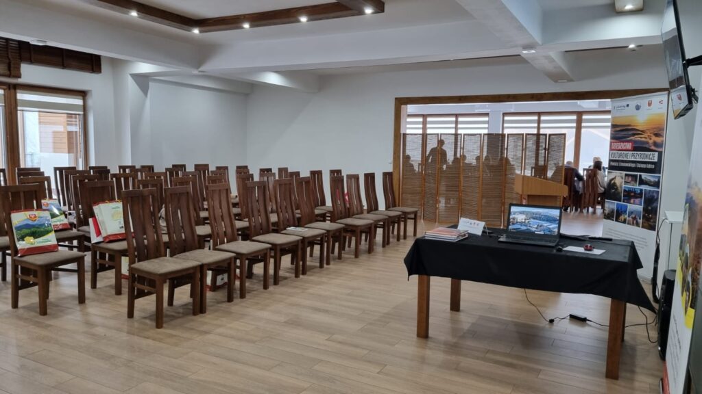 Wnętrze sali w hotelu Serce Beskidu przygotowane na oficjalne wydarzenie. Widoczne rzędy eleganckich drewnianych krzeseł w układzie teatralnym oraz stół prezydialny z laptopem. Na krzesłach znajdują się materiały promocyjne, co podkreśla, że to idealna sala konferencyjna w okolicach Krakowa na konferencje i prezentacje biznesowe.